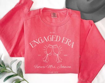 May include: A watermelon-colored crewneck sweatshirt featuring white screen-printed text that reads IN MY ENGAGED ERA, EST 2023, Future Mrs. Johnson, and I SAID YES! The design includes two clinking champagne flutes with ribbons.