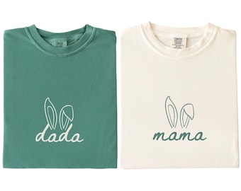 May include: A light green t-shirt with the word dada and white bunny ears printed on the front, and an ivory t-shirt with the word mama and green bunny ears printed on the front. Both shirts feature a crew neckline and Comfort Colors labels.