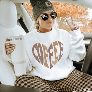 May include: White sweatshirt with a heart-shaped "COFFEE" design in shades of brown. The sweatshirt is worn with plaid pants and a gray beanie. A large white tumbler is held in the hand.