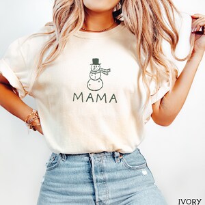 May include: An ivory short-sleeve t-shirt featuring a dark green line-art graphic of a snowman wearing a top hat and scarf, positioned above the text MAMA in a simple green font. The shirt has a crew neckline and a relaxed, casual fit.