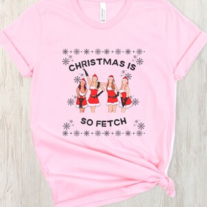 Peut inclure: T-shirt rose avec un motif de Noël représentant quatre femmes habillées en Père Noël avec le texte "Christmas is so fetch".