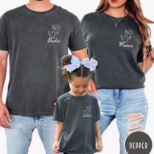 Op de afbeelding: Een familie draagt bijpassende donkergrijze t-shirts. Elk shirt heeft een witte ooievaar met ballonnen. De shirts lezen "Dada", "Mama" en "Sister". Ze dragen blauwe jeans.