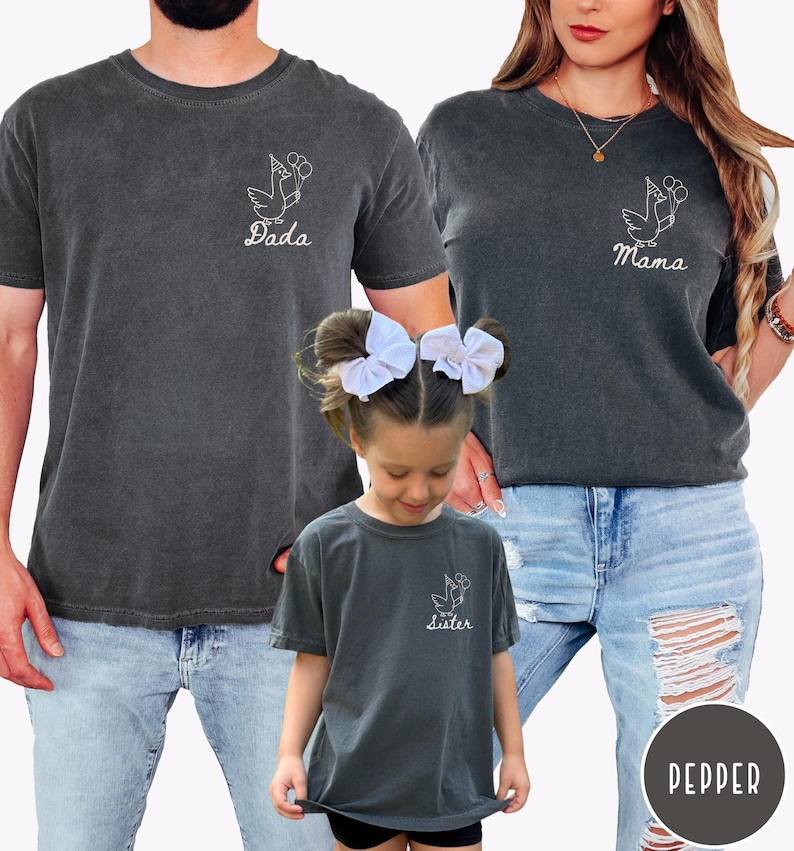Op de afbeelding: Een familie draagt bijpassende donkergrijze t-shirts. Elk shirt heeft een witte ooievaar met ballonnen. De shirts lezen "Dada", "Mama" en "Sister". Ze dragen blauwe jeans.