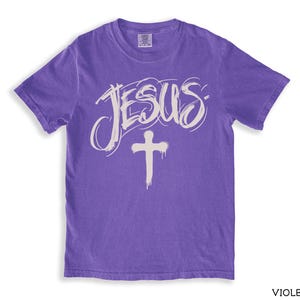 May include: A violet-colored t-shirt with the word "JESUS" in a handwritten style and a white cross graphic. The shirt has short sleeves and a crew neck. The word "VIOLET" is printed in the bottom right corner.