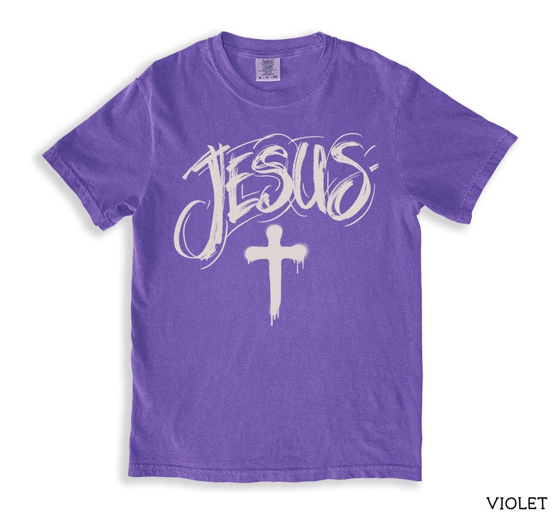 May include: A violet-colored t-shirt with the word "JESUS" in a handwritten style and a white cross graphic. The shirt has short sleeves and a crew neck. The word "VIOLET" is printed in the bottom right corner.
