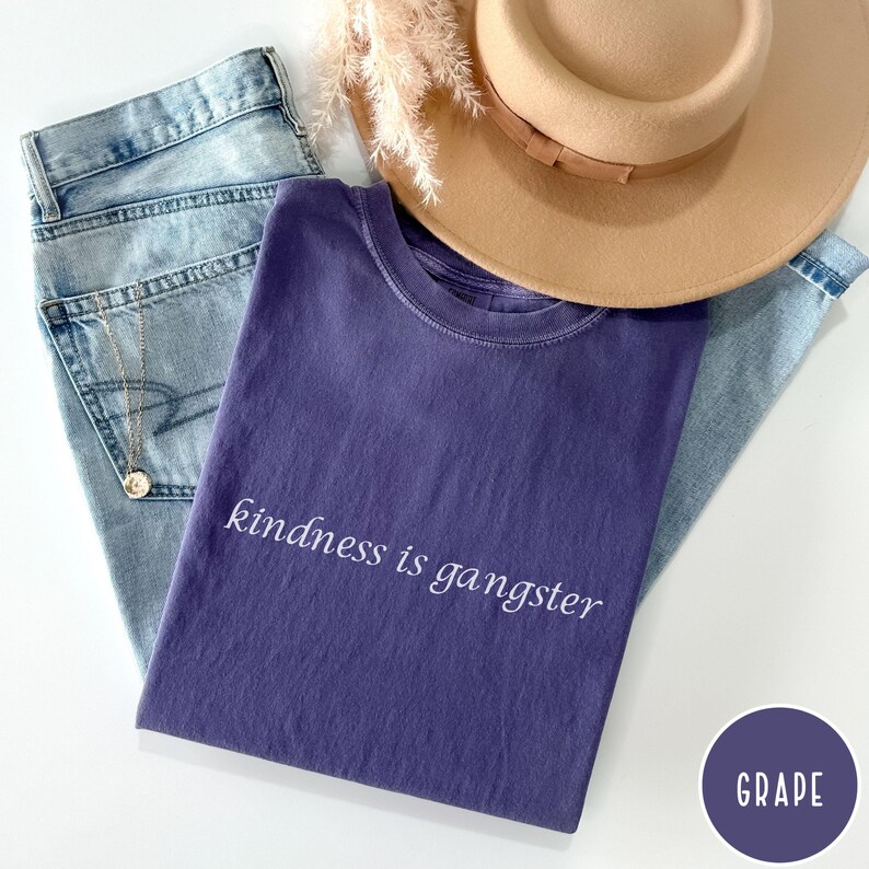 May include: A flat lay featuring a purple t-shirt with the phrase "kindness is gangster" in white script. The shirt is paired with light wash jeans, a tan hat, and a gold necklace. A purple circle with the word "GRAPE" is in the corner.