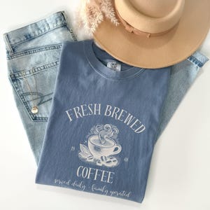 Op de afbeelding: Een blauw T-shirt met korte mouwen en een witte afbeelding van een dampende koffiekop met de tekst "FRESH BREWED COFFEE", "1968" en "served daily - family operated". Het shirt ligt op een lichtblauwe spijkerbroek en een beige hoed.
