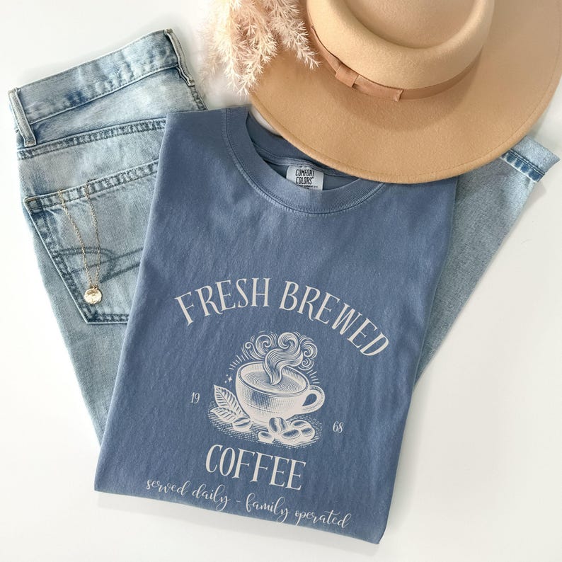 Op de afbeelding: Een blauw T-shirt met korte mouwen en een witte afbeelding van een dampende koffiekop met de tekst "FRESH BREWED COFFEE", "1968" en "served daily - family operated". Het shirt ligt op een lichtblauwe spijkerbroek en een beige hoed.