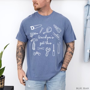 Peut inclure: T-shirt bleu jean avec un motif blanc d'outils divers, dont une scie, un marteau et une cl&eacute;. Le texte "Grandpa's got this" est centr&eacute; sous les outils. Le t-shirt est de style d&eacute;contract&eacute; &agrave; manches courtes.