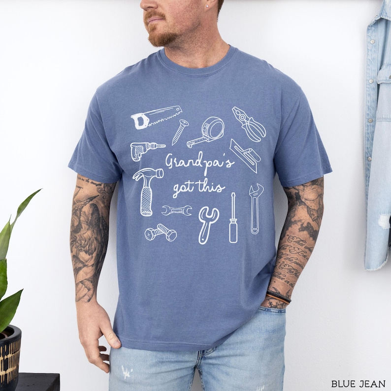 Peut inclure: T-shirt bleu jean avec un motif blanc d'outils divers, dont une scie, un marteau et une cl&eacute;. Le texte "Grandpa's got this" est centr&eacute; sous les outils. Le t-shirt est de style d&eacute;contract&eacute; &agrave; manches courtes.