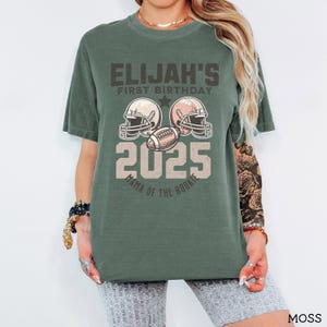 Puede incluir: Una camiseta de manga corta color verde musgo con un gr&aacute;fico de dos cascos de f&uacute;tbol americano, un bal&oacute;n de f&uacute;tbol y el texto ELIJAH'S FIRST BIRTHDAY 2025 MAMA OF THE ROOKIE. El dise&ntilde;o est&aacute; impreso en una paleta de colores vintage en el frente.