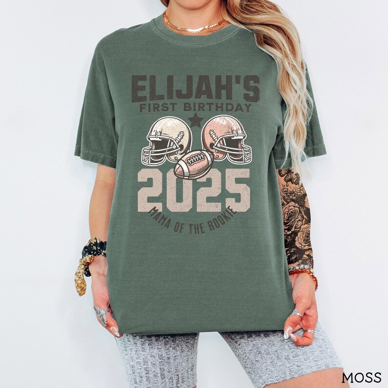 Puede incluir: Una camiseta de manga corta color verde musgo con un gr&aacute;fico de dos cascos de f&uacute;tbol americano, un bal&oacute;n de f&uacute;tbol y el texto ELIJAH'S FIRST BIRTHDAY 2025 MAMA OF THE ROOKIE. El dise&ntilde;o est&aacute; impreso en una paleta de colores vintage en el frente.