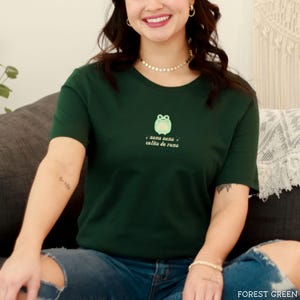 May include: A forest green t-shirt with a cartoon frog and the text "sana sana colita de rana" in a light pink font.
