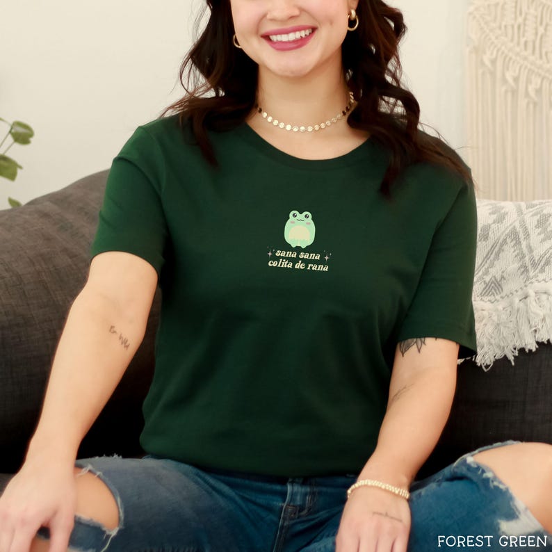 May include: A forest green t-shirt with a cartoon frog and the text "sana sana colita de rana" in a light pink font.