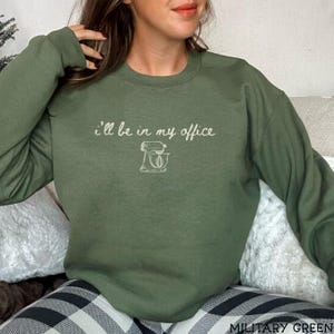 May include: An olive green crewneck sweatshirt featuring the white text "I'll be in my office" above a simple line drawing of a stand mixer. The garment has long sleeves and a ribbed neckline, providing a casual look for everyday wear.