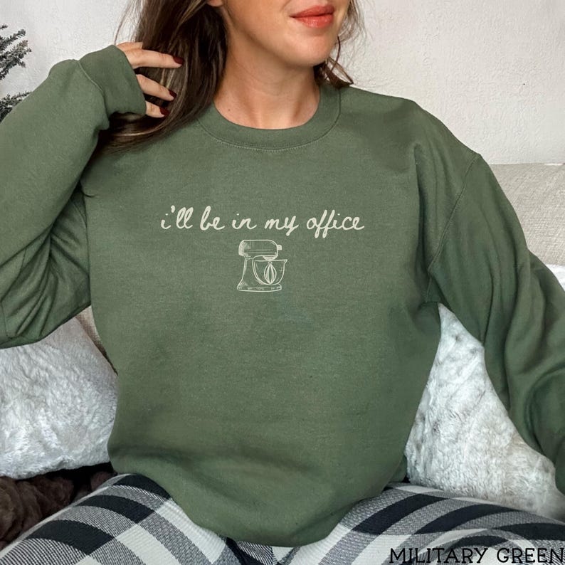 May include: An olive green crewneck sweatshirt featuring the white text "I'll be in my office" above a simple line drawing of a stand mixer. The garment has long sleeves and a ribbed neckline, providing a casual look for everyday wear.