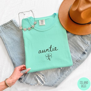 May include: A light teal t-shirt with the word "auntie" in black script and a small shamrock graphic. The shirt is laid on top of a pair of distressed blue jeans, with a brown hat and a floral necklace. The shirt is labeled "L-G".