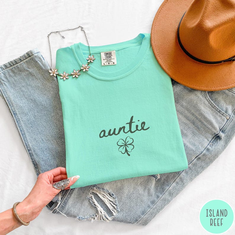 May include: A light teal t-shirt with the word "auntie" in black script and a small shamrock graphic. The shirt is laid on top of a pair of distressed blue jeans, with a brown hat and a floral necklace. The shirt is labeled "L-G".