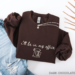May include: A dark chocolate brown crewneck sweatshirt featuring the white text "i'll be in my office" in a cursive script font above a line-art illustration of a stand mixer. The garment is paired with light blue distressed denim jeans.