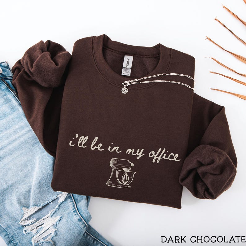 May include: A dark chocolate brown crewneck sweatshirt featuring the white text "i'll be in my office" in a cursive script font above a line-art illustration of a stand mixer. The garment is paired with light blue distressed denim jeans.