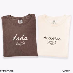 以下が含まれることがあります： エスプレッソブラウンとアイボリーのクルーネックTシャツ2枚。エスプレッソのシャツには白い筆記体で「dada」という文字と小さな飛行機のグラフィックが描かれています。アイボリーのシャツにはダークブラウンの筆記体で「mama」という文字と飛行機のグラフィックが描かれています。