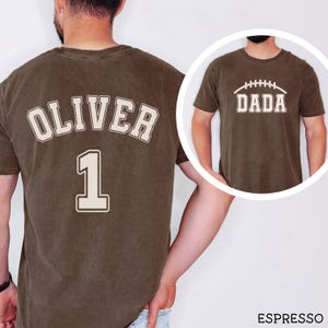 May include: An espresso-colored short-sleeve t-shirt featuring a cream-colored graphic on the back with the name OLIVER and the number 1 in a varsity font. The front view, shown in a circular inset, displays the word DADA with a football graphic.
