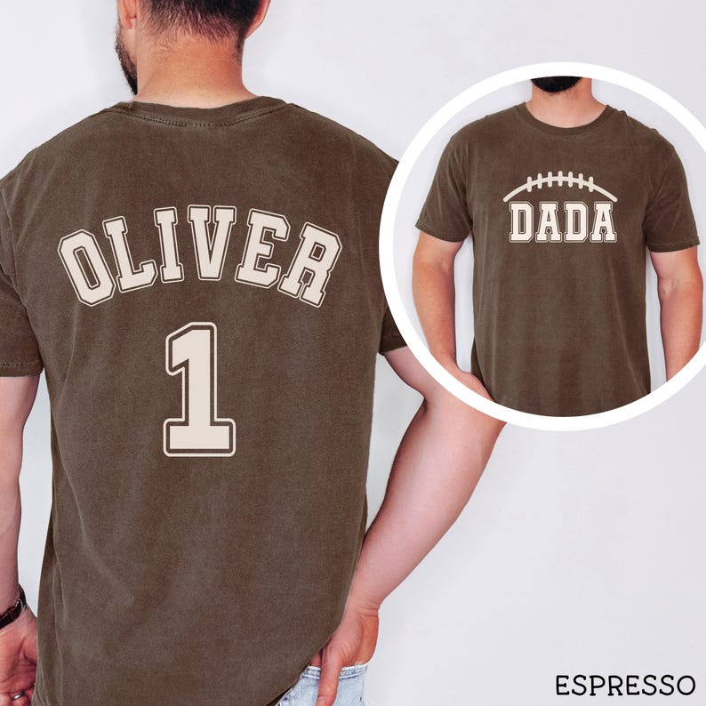 May include: An espresso-colored short-sleeve t-shirt featuring a cream-colored graphic on the back with the name OLIVER and the number 1 in a varsity font. The front view, shown in a circular inset, displays the word DADA with a football graphic.