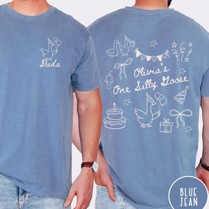 Op de afbeelding: Blauw spijkerbroekkleurig T-shirt met witte afbeelding van een gans met ballonnen en het woord "Dada". De achterkant van het shirt heeft een verjaardagsthema met de tekst "Olivia's One Silly Goose" en andere feestelijke illustraties.