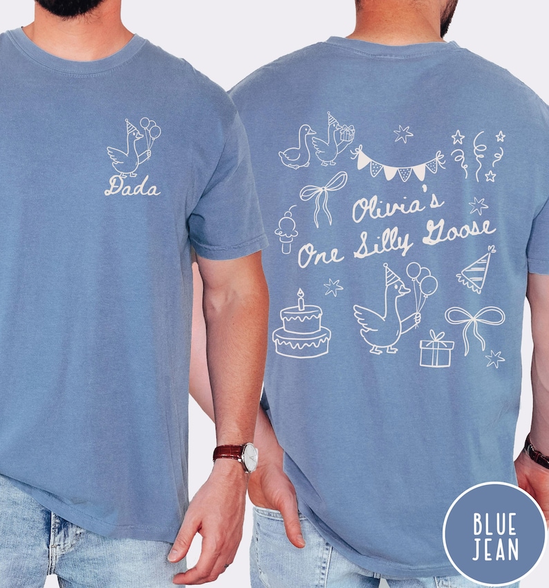 Op de afbeelding: Blauw spijkerbroekkleurig T-shirt met witte afbeelding van een gans met ballonnen en het woord "Dada". De achterkant van het shirt heeft een verjaardagsthema met de tekst "Olivia's One Silly Goose" en andere feestelijke illustraties.
