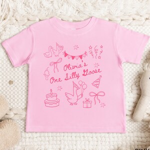 Op de afbeelding: Roze peuter-T-shirt met een grillig roze ontwerp. Het ontwerp bevat een gans met ballonnen, een verjaardagstaart, feesthoeden, strikken en de tekst "Olivia's One Silly Goose".