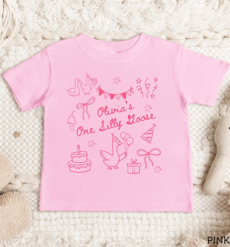 Op de afbeelding: Roze peuter-T-shirt met een grillig roze ontwerp. Het ontwerp bevat een gans met ballonnen, een verjaardagstaart, feesthoeden, strikken en de tekst "Olivia's One Silly Goose".