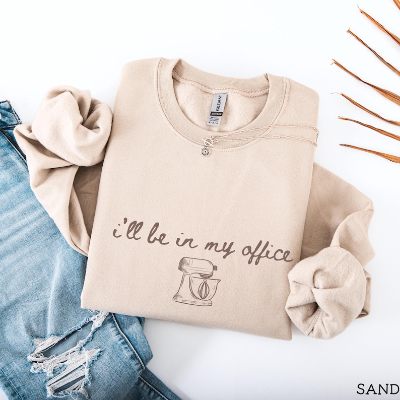 May include: A sand-colored crewneck sweatshirt featuring the dark brown text "I'll be in my office" above a line-art illustration of a stand mixer. The garment has long sleeves with ribbed cuffs and a ribbed neckline, paired with blue denim jeans.