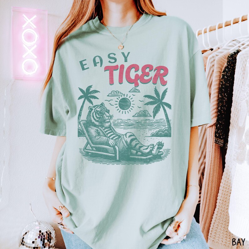 May include: A light blue t-shirt with the text "Easy Tiger" in pink and red lettering. A cartoon tiger wearing sunglasses is lounging on a beach chair with palm trees and a sun in the background.