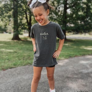 May include: A young girl wearing a dark grey t-shirt with the word "sister" and a drawing of a paintbrush and palette on it. She is smiling and looking down.