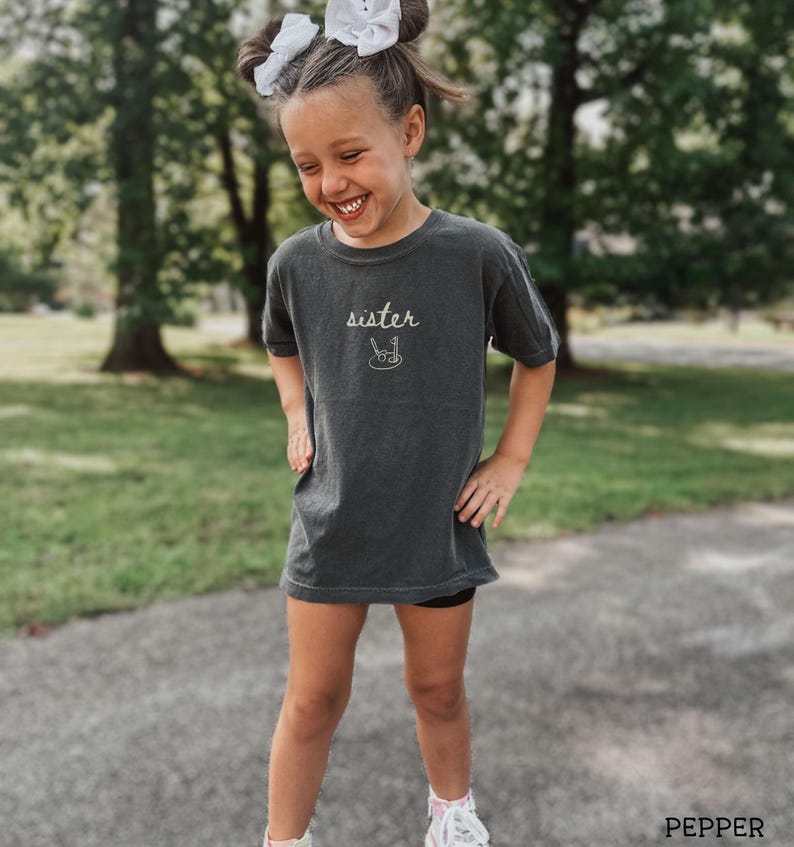 May include: A young girl wearing a dark grey t-shirt with the word "sister" and a drawing of a paintbrush and palette on it. She is smiling and looking down.