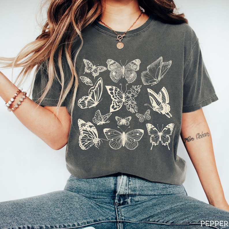 May include: A dark gray t-shirt with a vintage-style print of multiple butterflies in cream color. The butterflies are arranged in a random pattern across the front of the shirt.