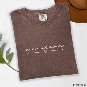 May include: A brown t-shirt with the word "mexicana" written in white script with a small heart in between two lines. The shirt is made of a soft, comfortable fabric.
