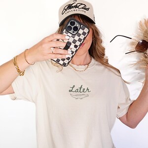 Pu&ograve; includere: T-shirt color avorio con la scritta "Later" e un disegno di coccodrillo. L'immagine mostra anche una custodia per telefono a scacchi bianchi e neri, un berretto da baseball beige con testo nero e occhiali da sole.