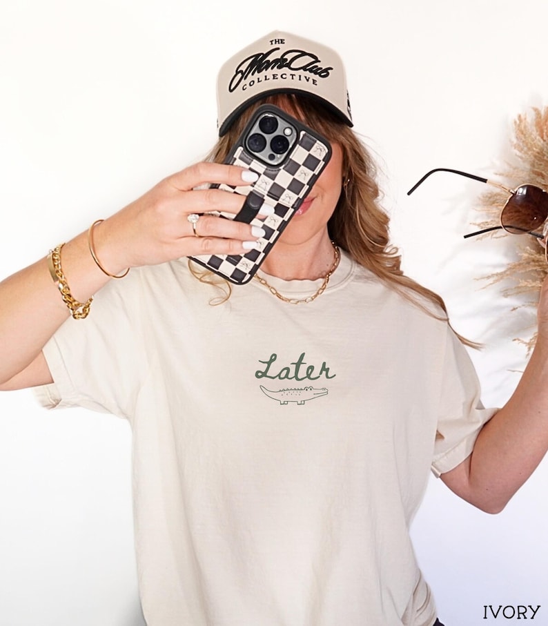 Pu&ograve; includere: T-shirt color avorio con la scritta "Later" e un disegno di coccodrillo. L'immagine mostra anche una custodia per telefono a scacchi bianchi e neri, un berretto da baseball beige con testo nero e occhiali da sole.