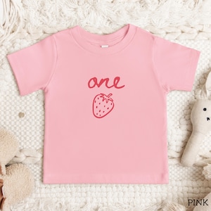 May include: A light pink short-sleeved t-shirt featuring the word one written in a dark pink cursive script above a dark pink line-art illustration of a strawberry. The shirt has a crew neckline and a simple, straight-cut hem design.
