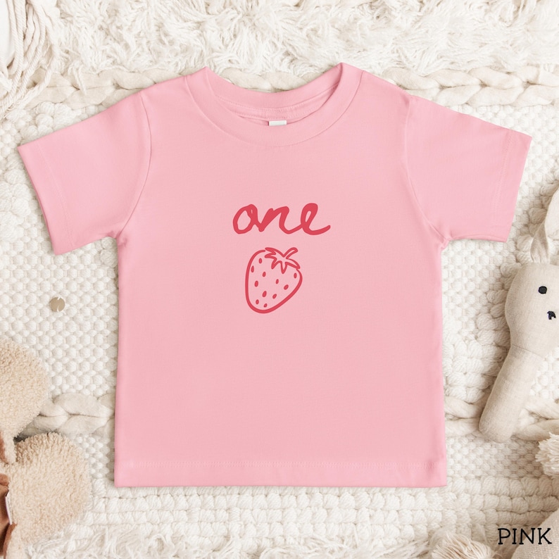 May include: A light pink short-sleeved t-shirt featuring the word one written in a dark pink cursive script above a dark pink line-art illustration of a strawberry. The shirt has a crew neckline and a simple, straight-cut hem design.