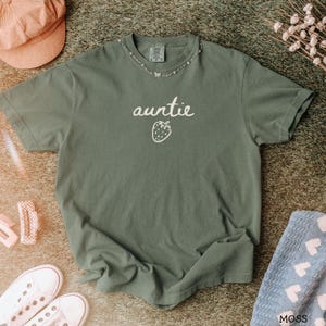 May include: A moss green short-sleeve t-shirt featuring the word "auntie" in white cursive script above a simple white line-art strawberry graphic. The shirt has a crew neckline and a visible brand tag at the inner collar area.