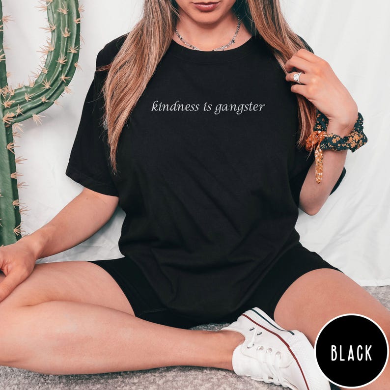 May include: Black t-shirt with the phrase "kindness is gangster" in a white, cursive font. The shirt is paired with black shorts and white sneakers. A black circle with the word "BLACK" is in the lower right corner.