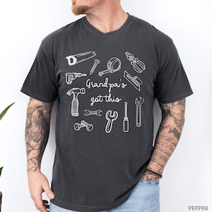 Peut inclure: T-shirt gris fonc&eacute; &agrave; col en V avec un motif blanc d'outils divers, dont une scie, un marteau et une cl&eacute;. Le texte "Grandpa's got this" est imprim&eacute; en blanc. Le t-shirt est en mati&egrave;re souple.