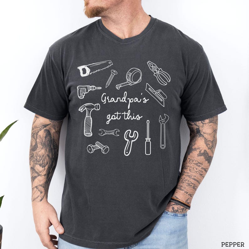 Peut inclure: T-shirt gris fonc&eacute; &agrave; col en V avec un motif blanc d'outils divers, dont une scie, un marteau et une cl&eacute;. Le texte "Grandpa's got this" est imprim&eacute; en blanc. Le t-shirt est en mati&egrave;re souple.