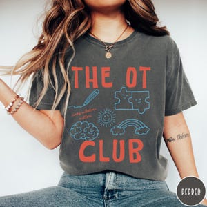 Peut inclure: Un t-shirt gris anthracite &agrave; manches courtes avec le texte rouge THE OT CLUB. Sous le texte se trouvent des dessins au trait bleu repr&eacute;sentant un crayon, deux pi&egrave;ces de puzzle, un soleil, un cerveau, un arc-en-ciel et le texte every milestone matters.