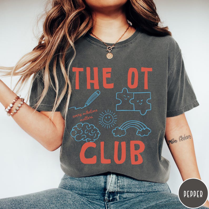 Peut inclure: Un t-shirt gris anthracite &agrave; manches courtes avec le texte rouge THE OT CLUB. Sous le texte se trouvent des dessins au trait bleu repr&eacute;sentant un crayon, deux pi&egrave;ces de puzzle, un soleil, un cerveau, un arc-en-ciel et le texte every milestone matters.