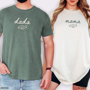 以下が含まれることがあります： モスグリーンのTシャツには白い文字でdadaと小さな飛行機のグラフィックが、アイボリーのTシャツにはダークグリーンの文字でmamaと小さな飛行機のグラフィックがプリントされています。どちらのシャツもクルーネックで半袖のカジュアルなデザインです。