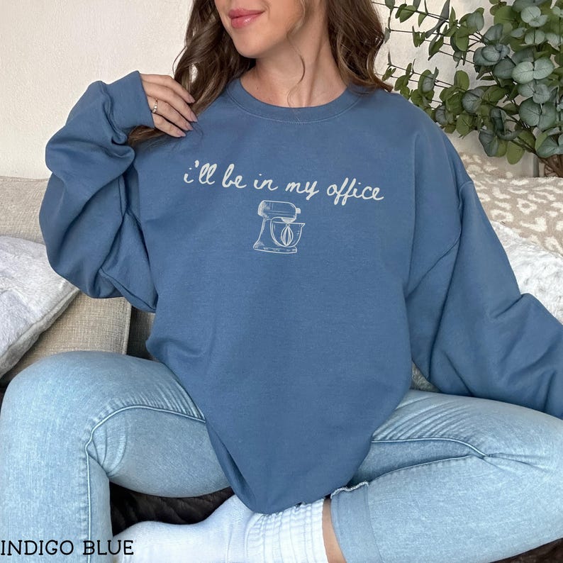 May include: An indigo blue crewneck sweatshirt featuring the white text "I'll be in my office" above a line-art illustration of a stand mixer. The long-sleeved garment has a ribbed collar, cuffs, and hem, providing a simple and functional design.