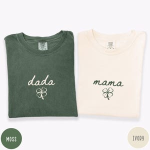 May include: Two t-shirts, one moss green and one ivory, are displayed. The moss green shirt has the word "dada" and a shamrock graphic. The ivory shirt has the word "mama" and a shamrock graphic. Both shirts are folded.